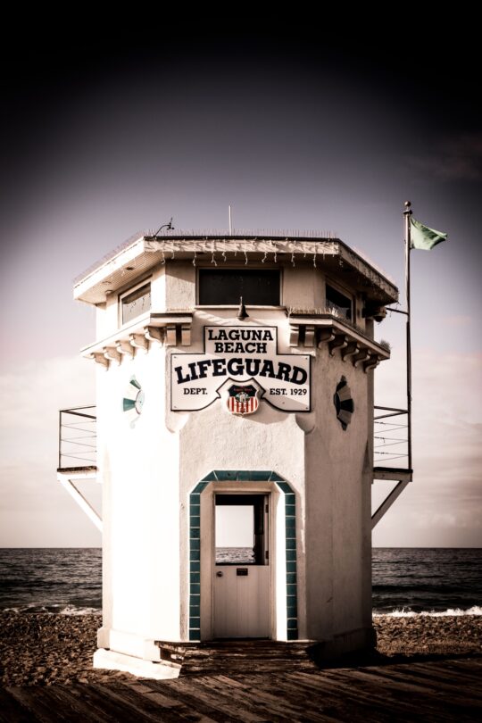 “Laguna Beach Lifeguard Tower at Main Beach, photographed head-on with the ocean behind it. The historic white tower features sea-blue tile accents around the door, a green flag flying on the right, and a sign reading ‘Laguna Beach Lifeguard Dept. Est. 1929.’ Dramatic sky and calm waves frame the iconic coastal landmark.