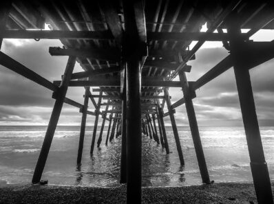 Black and white view beneath San Clemente Pier in Southern California, wooden pilings receding toward the Pacific under overcast skies.