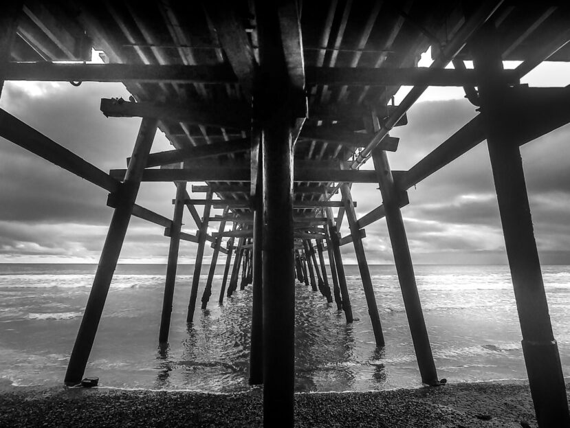 Black and white view beneath San Clemente Pier in Southern California, wooden pilings receding toward the Pacific under overcast skies.