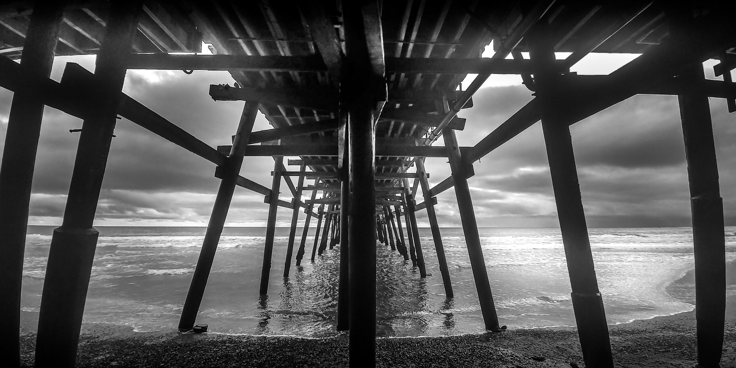 Black and white view beneath San Clemente Pier in Southern California, wooden pilings receding toward the Pacific under overcast skies.