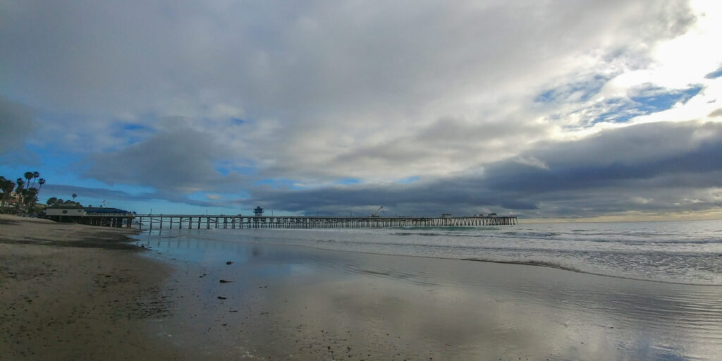 The San Clemente Pier stretches across the Pacific Ocean beneath layered winter clouds, reflected in wet sand along the shoreline.
