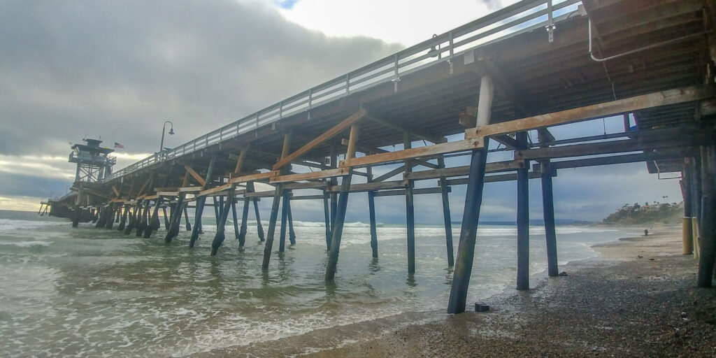 View from beneath the San Clemente Pier showing wooden pilings, rolling waves, and a cloudy sky along the shoreline.