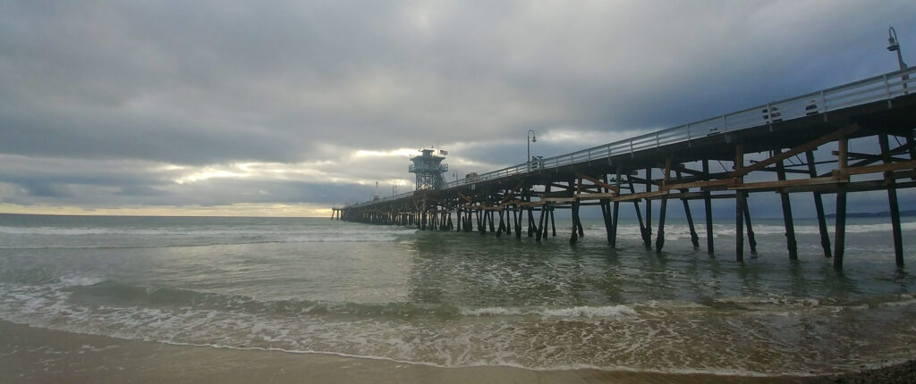The San Clemente Pier stretches over shallow waves beneath an overcast sky, with wooden pilings leading toward the horizon.