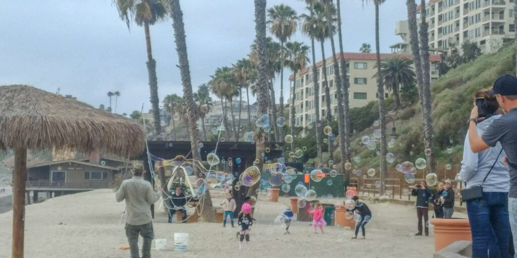 Children gather near the entrance to the San Clemente Pier as a street performer creates large floating bubbles on a crisp coastal afternoon.