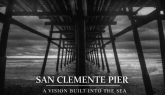 Black-and-white photograph taken beneath the San Clemente Pier, showing wooden pilings receding into the Pacific Ocean under a dramatic sky.