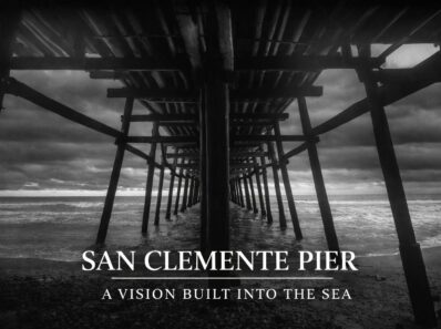 Black-and-white photograph taken beneath the San Clemente Pier, showing wooden pilings receding into the Pacific Ocean under a dramatic sky.