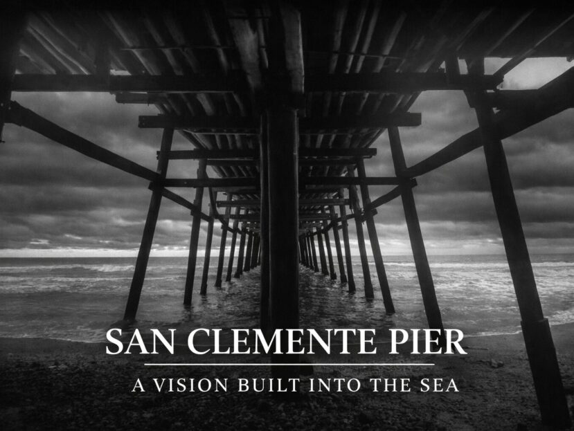 Black-and-white photograph taken beneath the San Clemente Pier, showing wooden pilings receding into the Pacific Ocean under a dramatic sky.