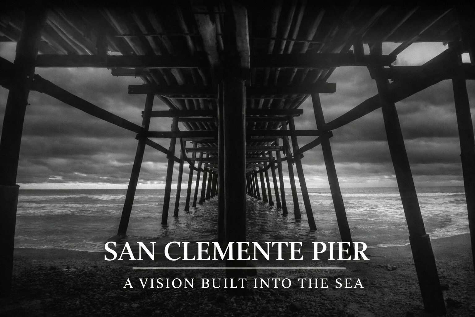 Black-and-white photograph taken beneath the San Clemente Pier, showing wooden pilings receding into the Pacific Ocean under a dramatic sky.