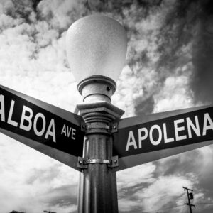 Black-and-white photo of a streetlamp with Balboa Ave and Apolena Ave signs pointing opposite directions, shot upward against a cloudy sky.