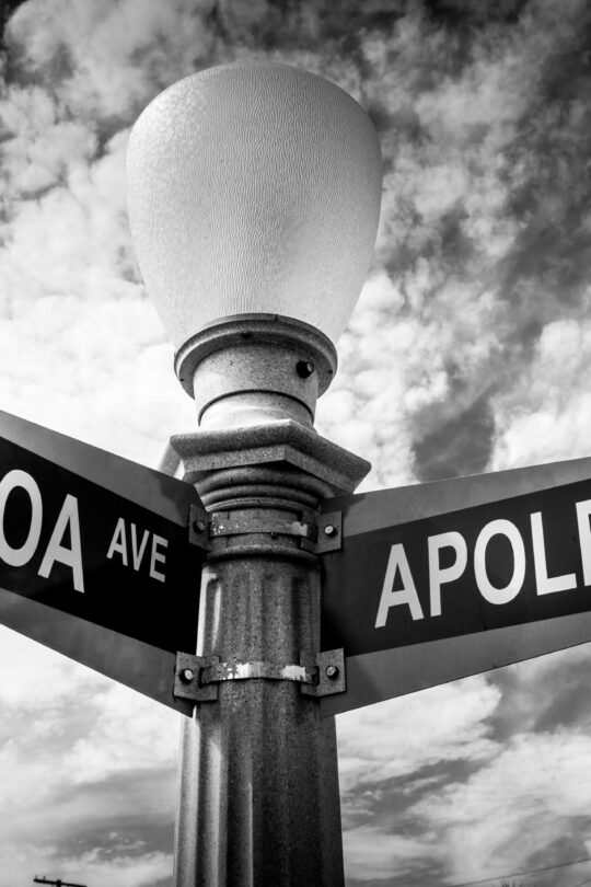 Black-and-white photo of a streetlamp with Balboa Ave and Apolena Ave signs pointing opposite directions, shot upward against a cloudy sky.