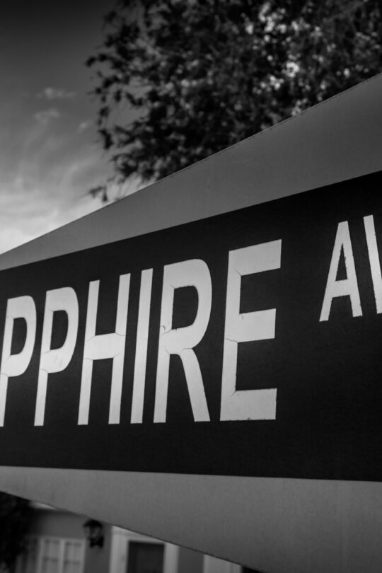 Black-and-white photograph of a Sapphire Ave street sign viewed at an angle, bold lettering against a cloudy sky with trees softly blurred in the background.