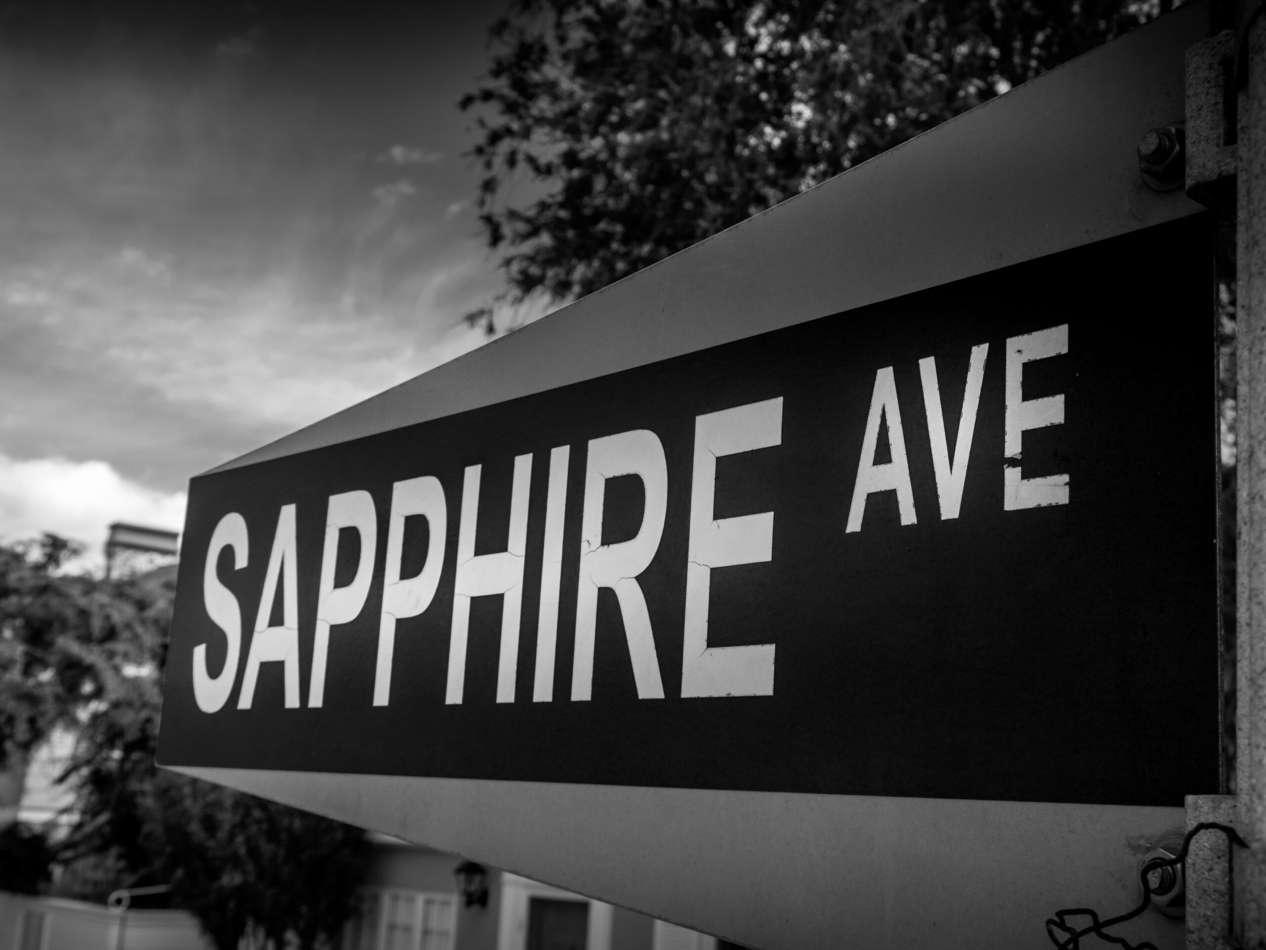 Black-and-white photograph of a Sapphire Ave street sign viewed at an angle, bold lettering against a cloudy sky with trees softly blurred in the background.